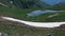 Travelers sit near tents on the shore of a mountain lake, and horses walk nearby.