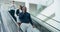 Traveling businessman talking on a phone, standing on an escalator in an airport. Corporate man traveling with luggage