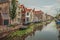 Tree-lined long canal with aquatic greenish plants, brick houses on its bank and cloudy day at Gouda.