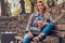 Trendy blonde female relaxes outdoor, drinking takeaway coffee while sitting on the bench in city park.