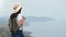 Trendy woman in hat with backpack posing on top of mountain over sea. Medium shot on RED camera
