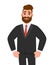 Trendy young bearded businessman standing and holding hands in pockets. Cheerful stylish person in black colour formal business.