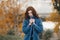 Trendy young woman in warm blue coat posing in the autumn park. She closing the coat by her hands