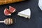 Triangular appetizing piece of camembert cheese, pieces of figs in honey and a wooden spoon for honey and baguette on a graphite b