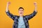 Triumphant young guy lifting arms above his head, shouting in excitement on orange studio background