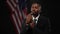 Troubled thoughtful African American elegant man in suit waiting for public speaking at black background with USA flag