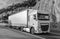 Truck carrying goods on a beautiful mountain road. The truck is white with the headlights on. Black and white photo