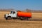 A truck for the transportation of gasoline and fuel with an orange tank rides in a yellow field on the road during the delivery of