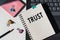 Trust word printed on notepad lying on laptop next to pink sticknotes, paper clips and black pencil