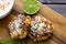 TTwo shrimp cakes on a cutting board with a lemon garlic dipping sauces lemon wedge and a stem or rosemary on a wooden table..