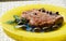 Tuna steak served with olives, basil and capper in yellow plate in blurry background