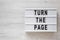 `Turn the page` words on a lightbox on a white wooden background, top view. Overhead, from above, flat lay. Copy space