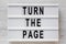 `Turn the page` words on a lightbox on a white wooden surface, top view. Overhead, from above, flat lay. Close-up