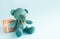 Turquoise soft teddy bear with an embroidered heart holds a gift box and a bow on a blue background. Children`s toy. Love, a gift