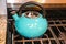 Turquoise water kettle on lit gas stove - textured handle and steel grills and marble countertop - Close-up and selective focus