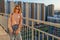 Tween redhead girl in pullover, jeans and sunglasses standing on balcony against high-rise multi-storey residential building at