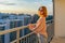 Tween redhead girl in pullover, jeans and sunglasses standing on balcony against high-rise multi-storey residential building at