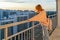 Tween redhead girl in pullover, jeans and sunglasses standing on balcony against high-rise multi-storey residential building at