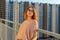 Tween redhead girl in pullover, jeans and sunglasses standing on balcony against high-rise multi-storey residential building at