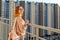 Tween redhead girl in pullover, jeans and sunglasses standing on balcony against high-rise multi-storey residential building at