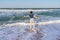 Tween slender girl standing on sand beach of sea coast against stormy waves in sunny summer day. Young woman spending summer