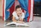 Tween thinking boy with curly hair in toy tent house lying and reading book at home