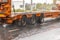 twelve wheels orange step deck trailer moving on wet road at rainy day with water splashes