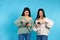 The twin girls are impressed while looking at smartphone, pointing at the screen. Blue background
