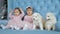 Twin kids in pink dresses and bows on head near white fluffy puppies sit on couch