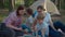 Two 30s mothers and two sons watching tablet computer laying on picnic blanket on summer camping vacation with tent in