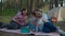 Two 30s mothers and two sons watching tablet computer laying on picnic blanket on summer camping vacation with tent in