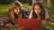 Two adolescent Caucasian girls lying on green park lawn in sunbeam talking surfing social media on laptop. Front view