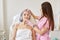 Two adorable women posing over white wall in beauty parlor, client laying on coach, having revitalizing cream under one eye, looks