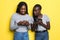 Two african young women spying each other while both using cell phones isolated over yellow background