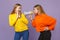 Two amazed shocked young blonde twins sisters girls in vivid colorful clothes scream on megaphone  on pastel