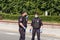 Two armed bodyguards in black uniforms, protective masks, gloves patrol city streets during global pandemic.