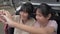 Two Asian adorable female teenagers enjoy making hand gesture heart shape together while sitting on rear side of hatchback car.
