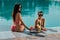 Two attractive girls in sunglasses and bikini drinking cocktails at poolside.