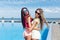 Two attractive girls in sunglasses with long hair are having fun near pool on the sun. Brunette girl wears short pink