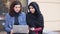 Two attractive young women sitting outside and using laptop. Cross-cultural friendship. Young muslim woman in black