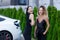 Two beautiful businesswomen in black outfut smiling and posing. Portrait of girls with laptop in front of electric car charging in