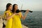 Two beautiful girls violinists in yellow concert dresses are playing electric violins with sunset, mountains and Mediterranean sea