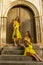 Two beautiful twin sisters violinists in yellow concert dresses are posing with electric violins near ancient wooden gate in old