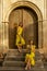 Two beautiful twin sisters violinists in yellow concert dresses are posing with electric violins near ancient wooden gate in old