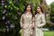 Two beautiful twins young women in trench coats near blooming lilac