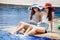 two beautiful Young Asian women in big summer hat and sunglasses sitting on the edge of the swimming pool with feet in water