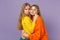 Two beautiful young blonde twins sisters girls in vivid colorful clothes hugging and looking camera isolated on pastel