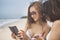 Two beautiful younger woman wearing bikini looking on smart phone beside vacation sea beach