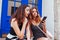 Two best female friends chatting while using smartphone in city. Happy girls talking and laughing