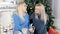 Two blond hair ladies speaking about something siting on the carpet near the Christmas tree